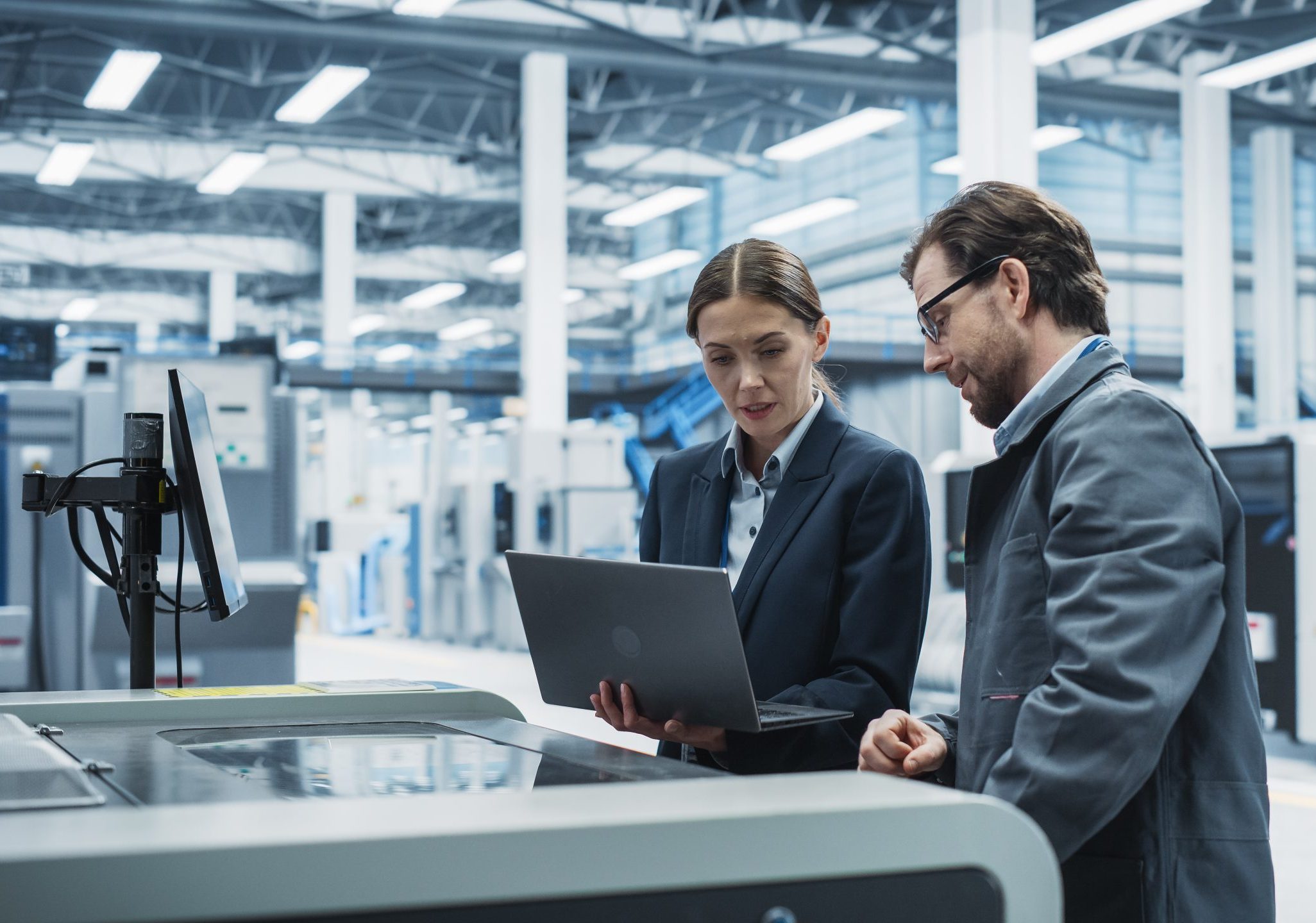 Caucasian Female Manager Using Laptop And Talking To Male Industrial Engineer At Modern Electronics Factory. Colleagues Using Soldering Jet Printer For Super Computer Motherboards Production.